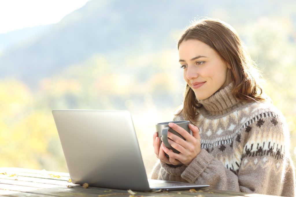 woman drinking coffee at picnic table looking at laptop